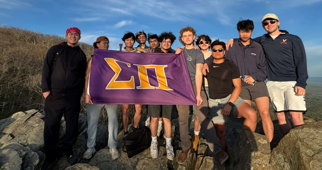 Group of actives at the summit of a mountain. They are proudly displaying the purple and yellow sigma pi flag.
